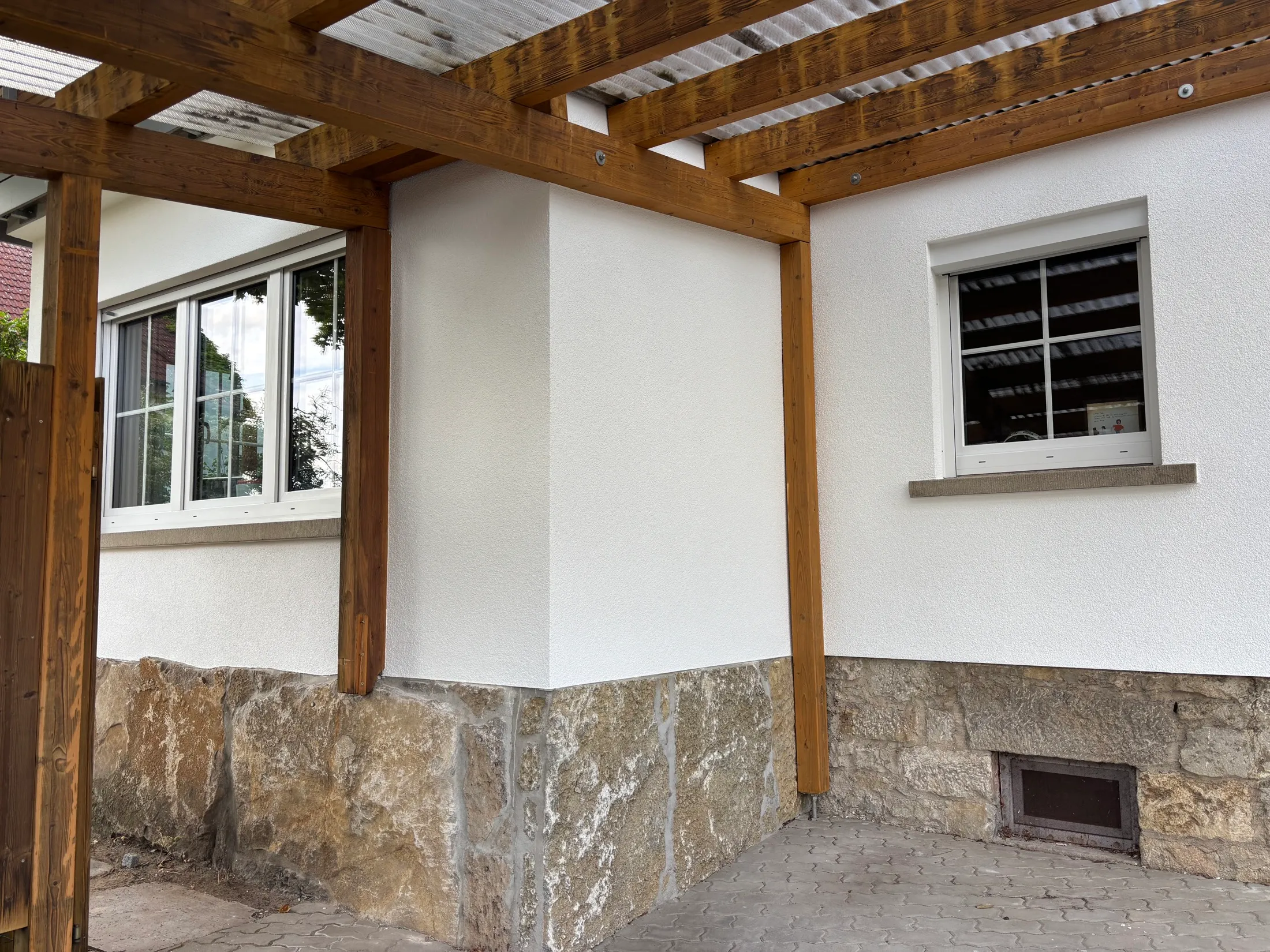 Fertig verputzte und gestrichene Gebäudeecke mit Natursteinsockel und Holzpergola